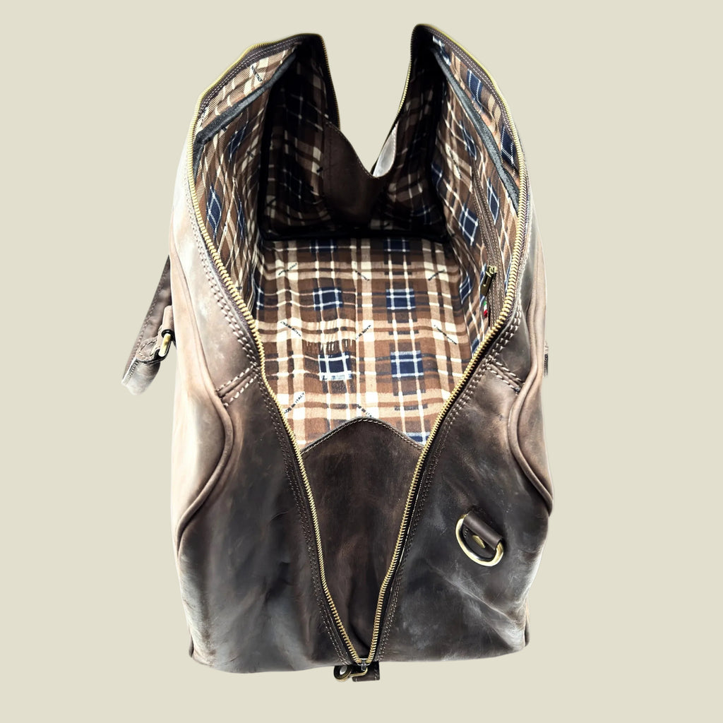 A dark brown distressed leather duffel bag shown open, revealing a brown, beige, and blue plaid patterned lining, set against a plain white background.