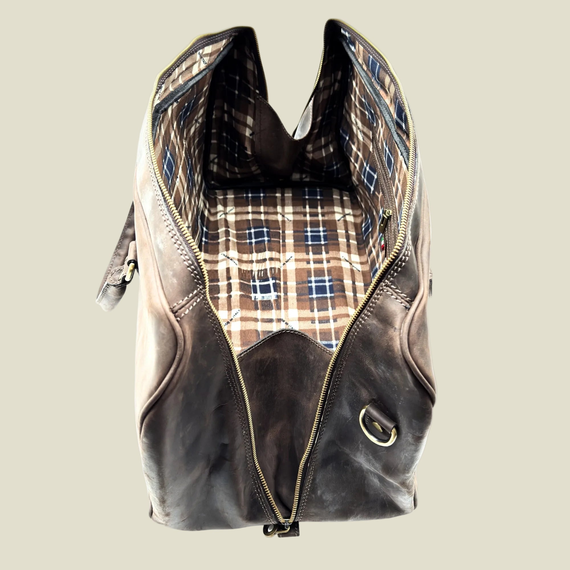 A dark brown distressed leather duffel bag shown open, revealing a brown, beige, and blue plaid patterned lining, set against a plain white background.