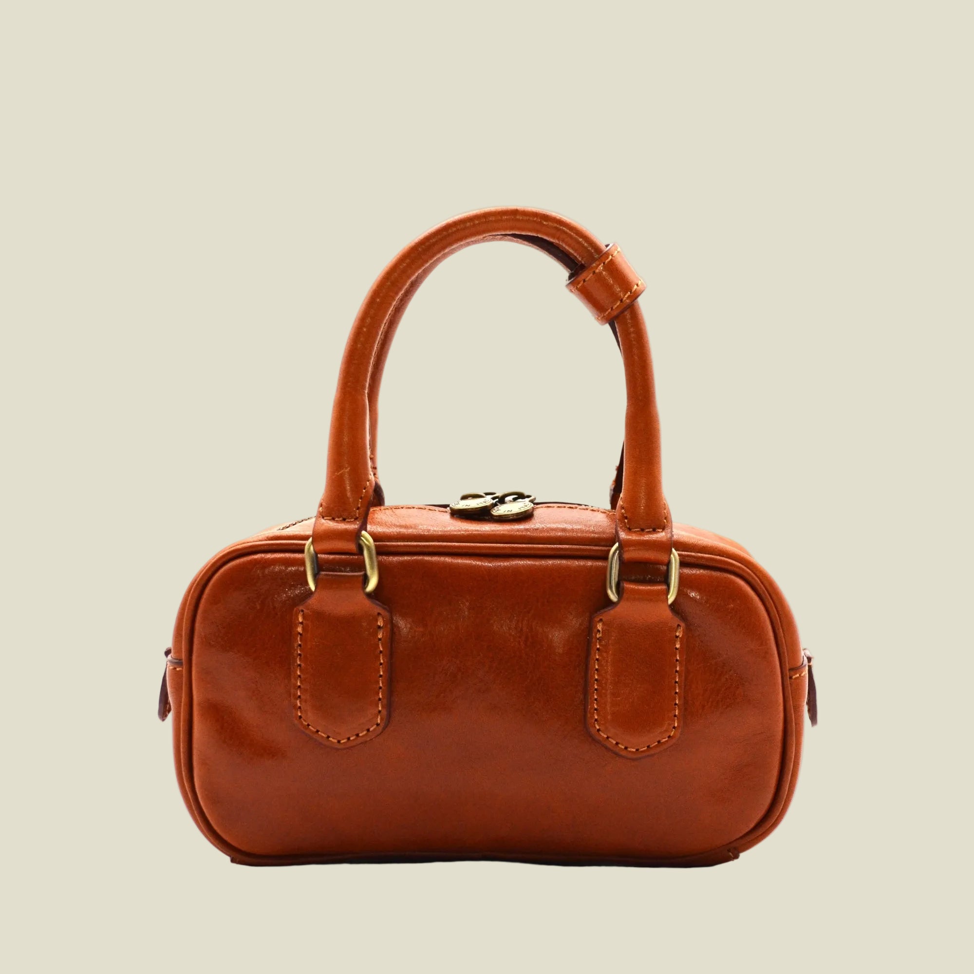 A small brown leather handbag with double top handles, brass-tone hardware, and a zip-top closure, photographed against a plain white background.