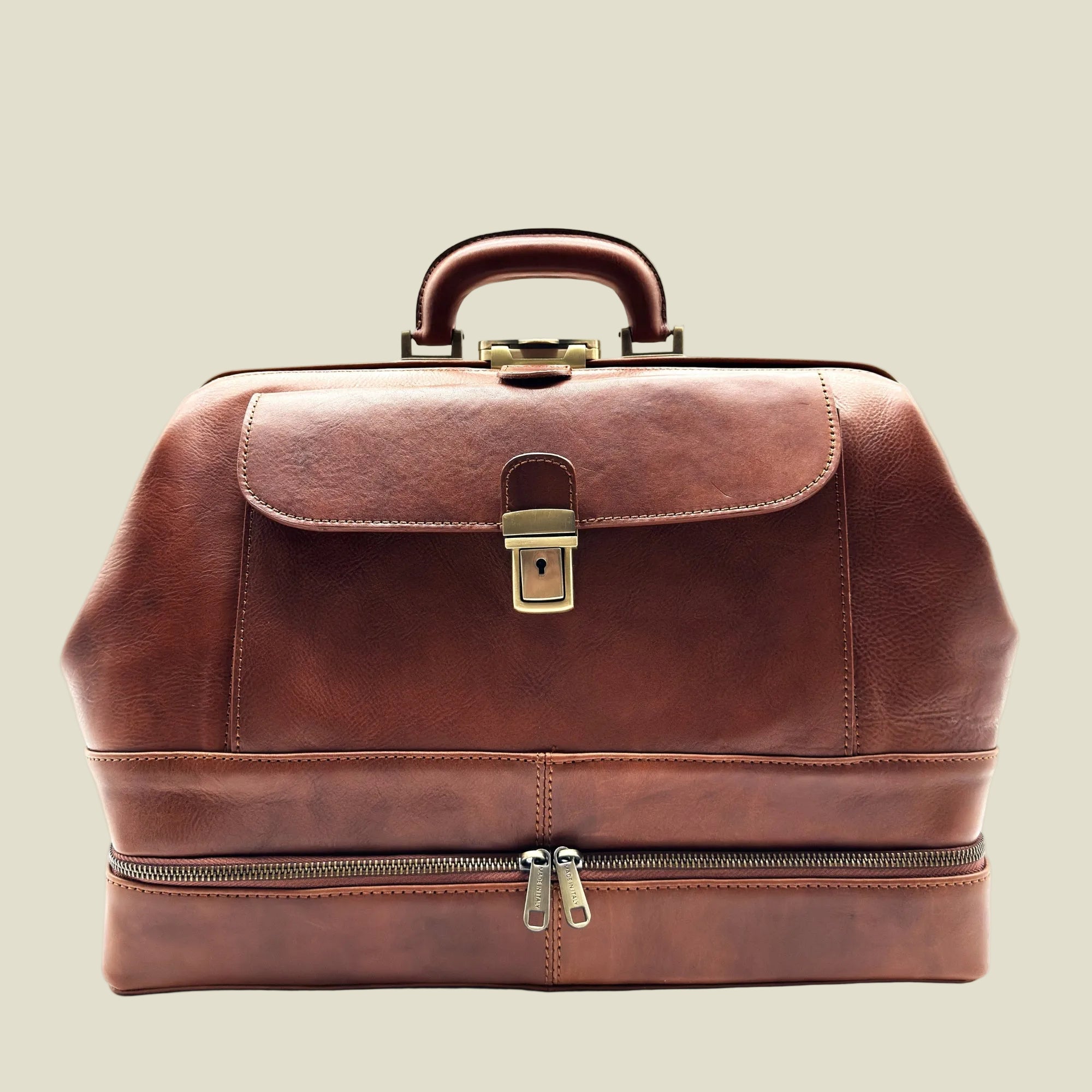 A brown leather doctor-style briefcase with a top handle, front flap, brass lock, and a zippered lower compartment, shown on a plain white background.