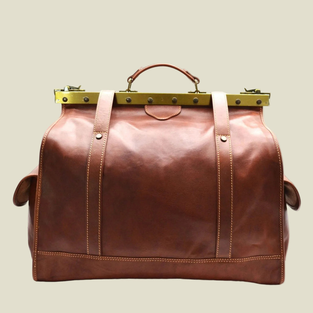 A brown leather duffel bag with brass hardware, featuring a top handle, vertical strap details, and side pockets, shown against a plain white background.