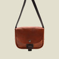 A brown leather crossbody bag with a dark brown strap and a snap-button closure on a plain white background.