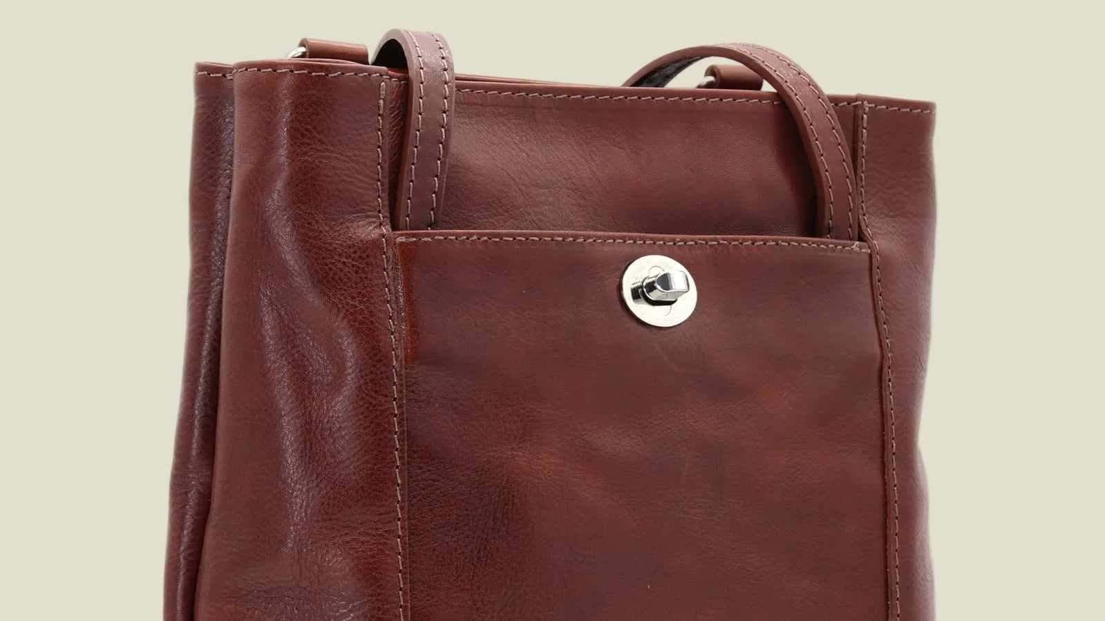A close-up of a brown leather tote bag with a front pocket and a silver-tone snap button, shown against a plain white background.