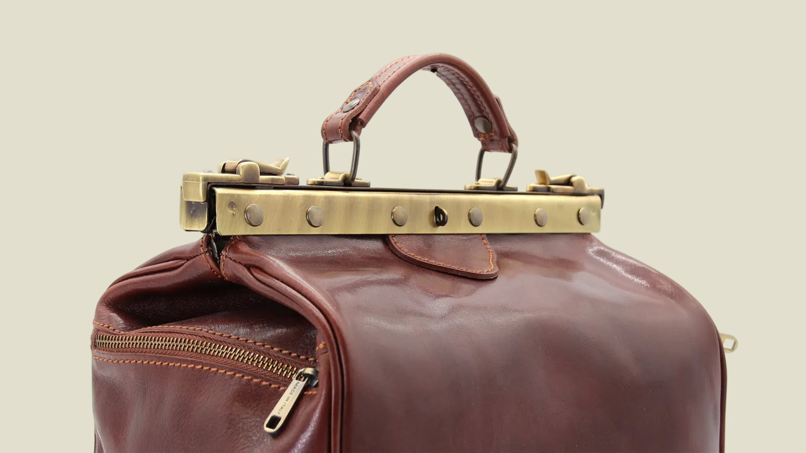 A close-up of a brown leather bag featuring a prominent brass-tone metal clasp closure across the top and a top handle, set against a plain white background.