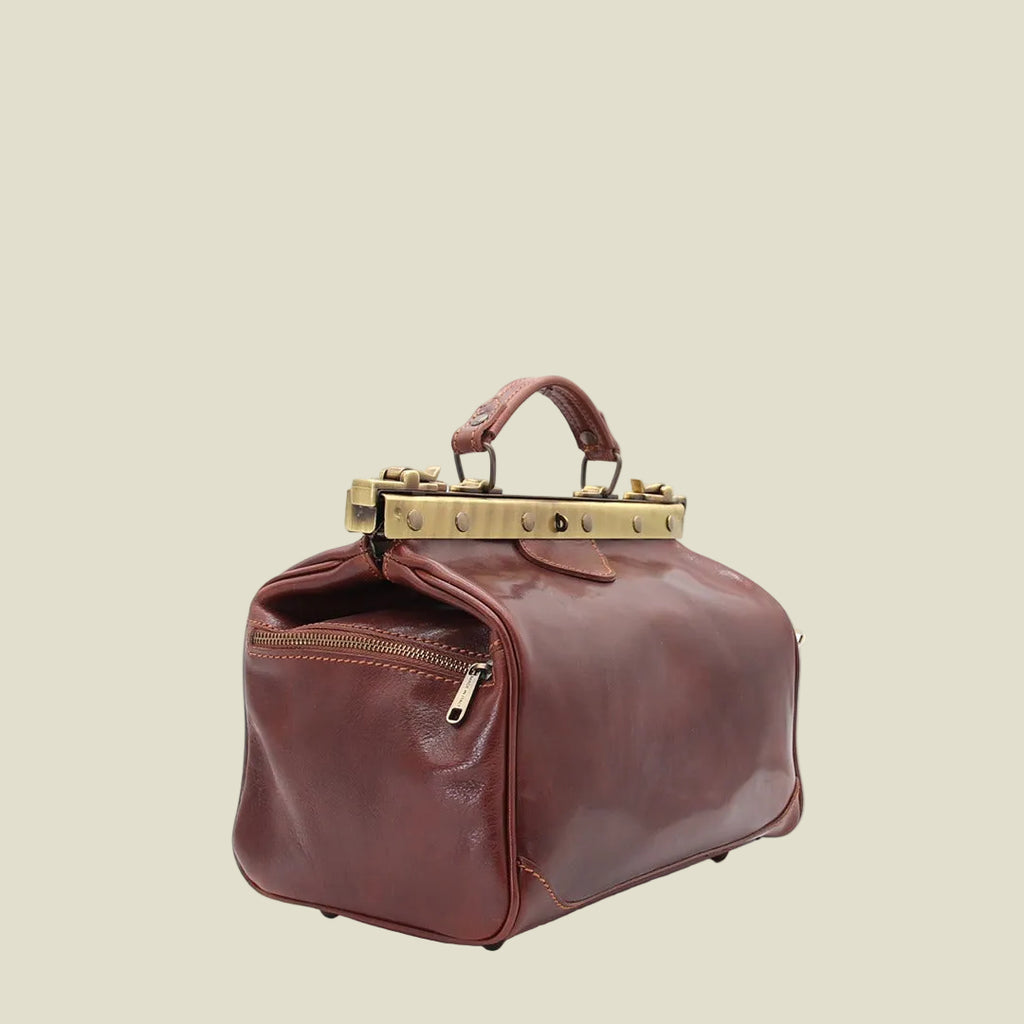A brown leather travel-style handbag with a top handle, a zippered front pocket, and a decorative gold-tone metal frame on top, shown on a plain white background.
