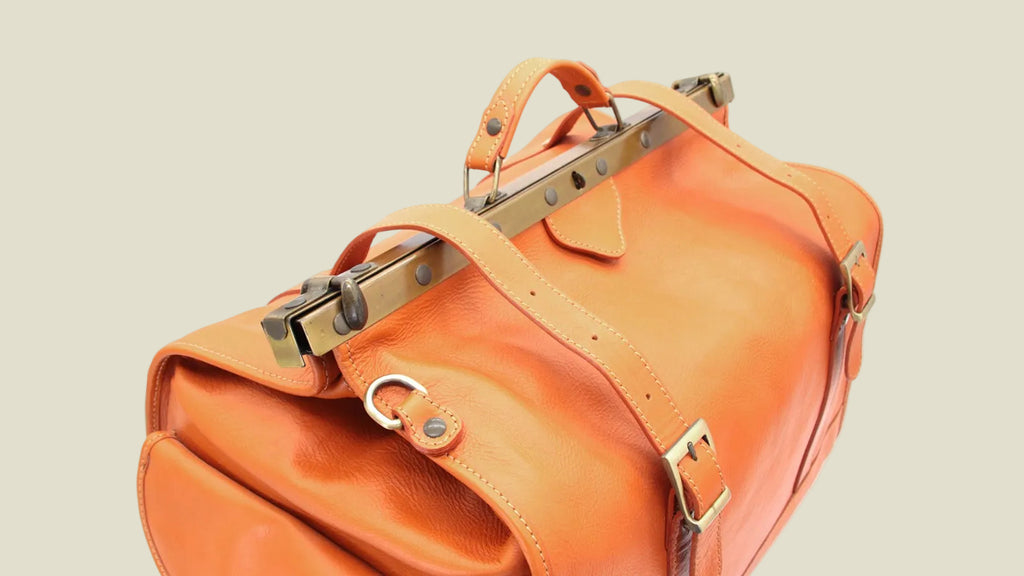 An orange leather travel duffel bag with brass hardware and top handles, shown on a plain white background.