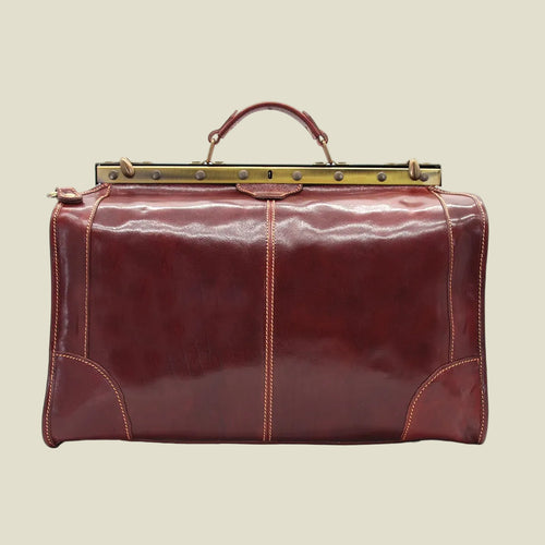 A vintage-style leather duffel bag in deep burgundy red, featuring a top handle, brass-toned metal frame, and contrast stitching.