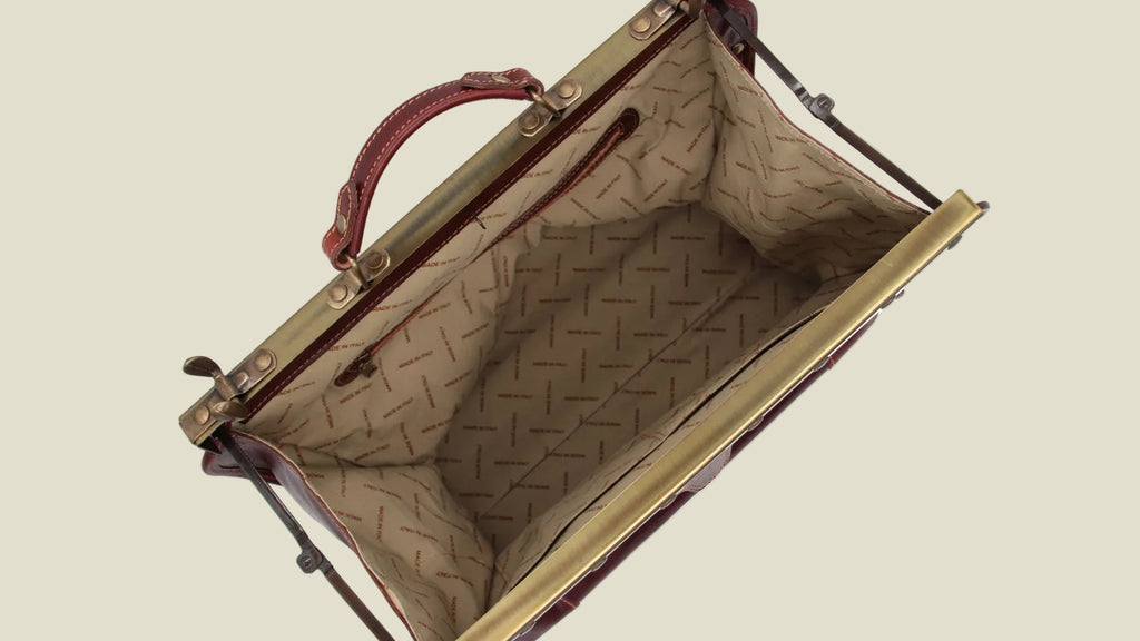 An open vintage-style clutch or handbag with a gold-tone metal frame, a brown leather handle, and a beige patterned interior lining, shown against a plain white background.
