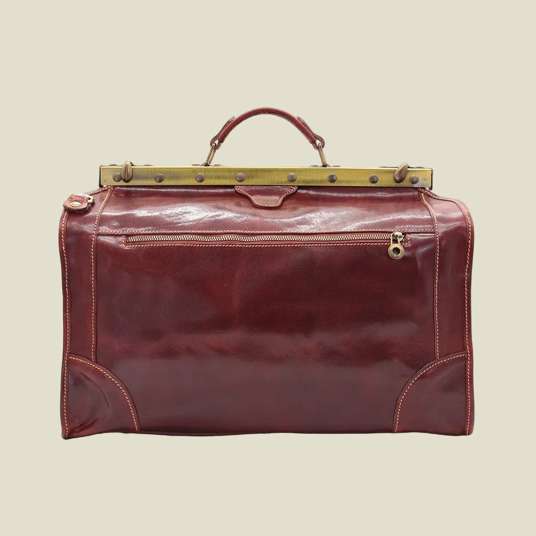 A vintage-style leather duffel bag in a glossy reddish-brown color, featuring a brass-toned metal frame across the top, a top handle, and a front zippered pocket.