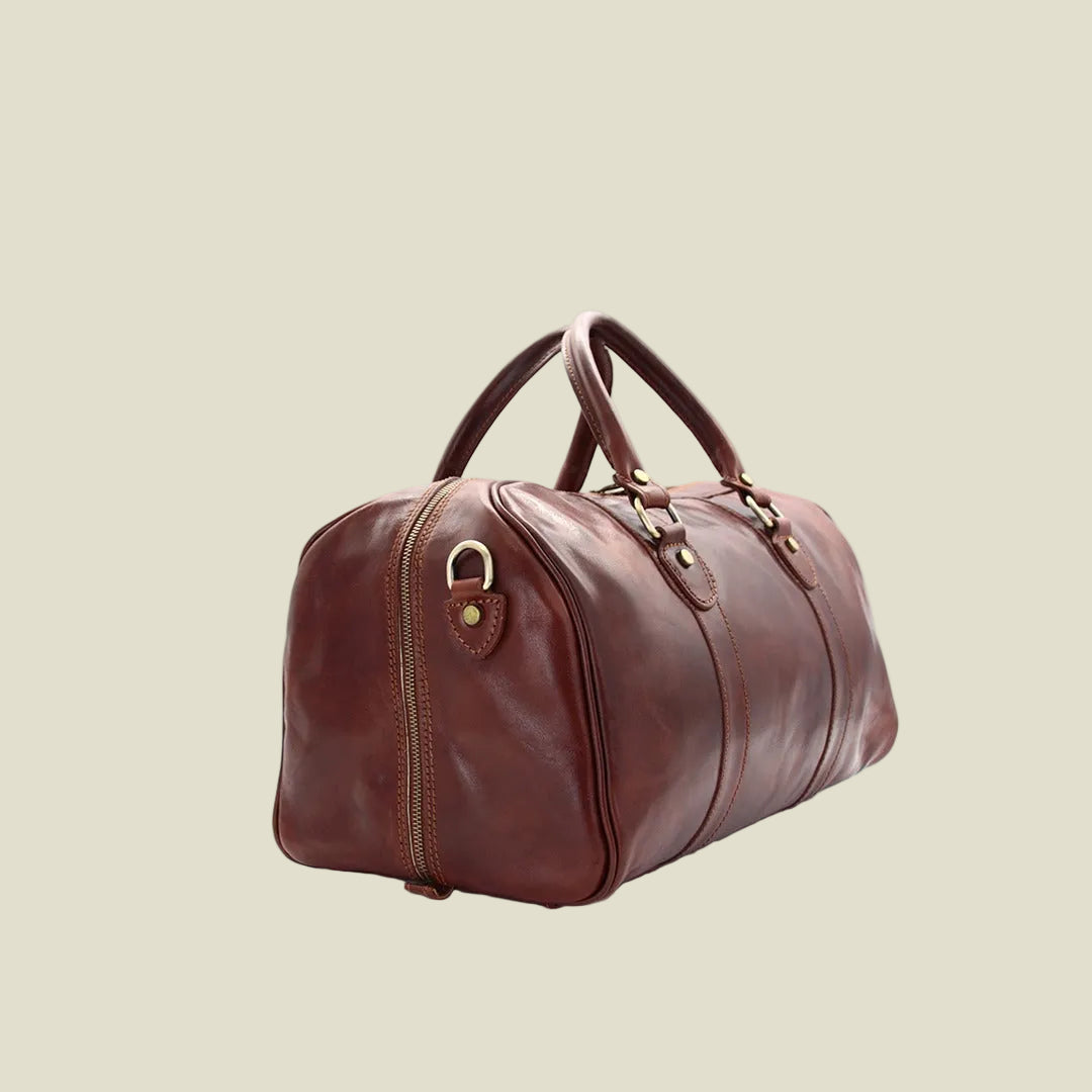 A brown leather duffel bag with dual top handles, a front zipper pocket, and brass-tone hardware, photographed on a clean white background.