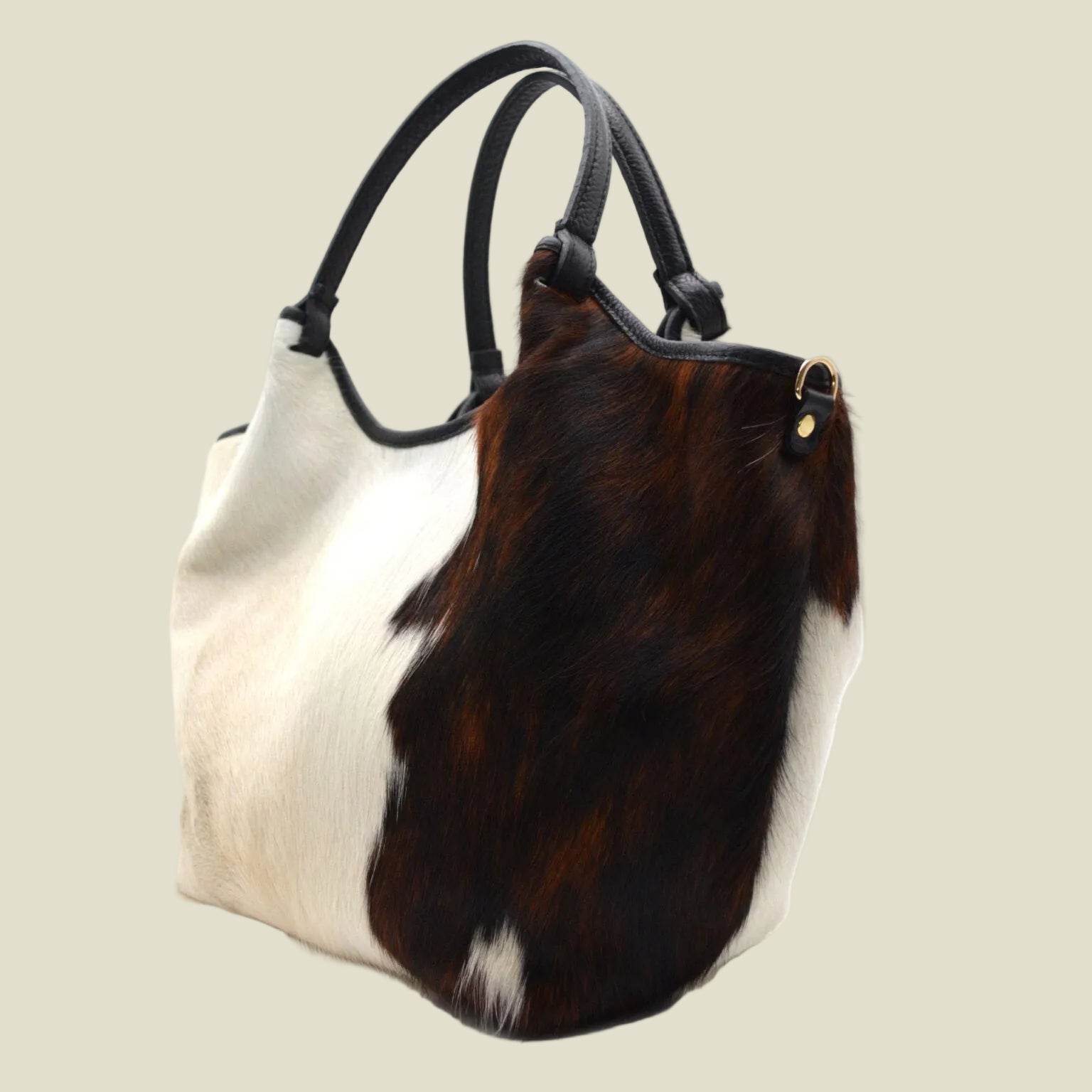 A cowhide shoulder bag with a split design of cream-white and dark brown hair-on-hide, featuring black leather handles and a small gold-tone hardware accent.