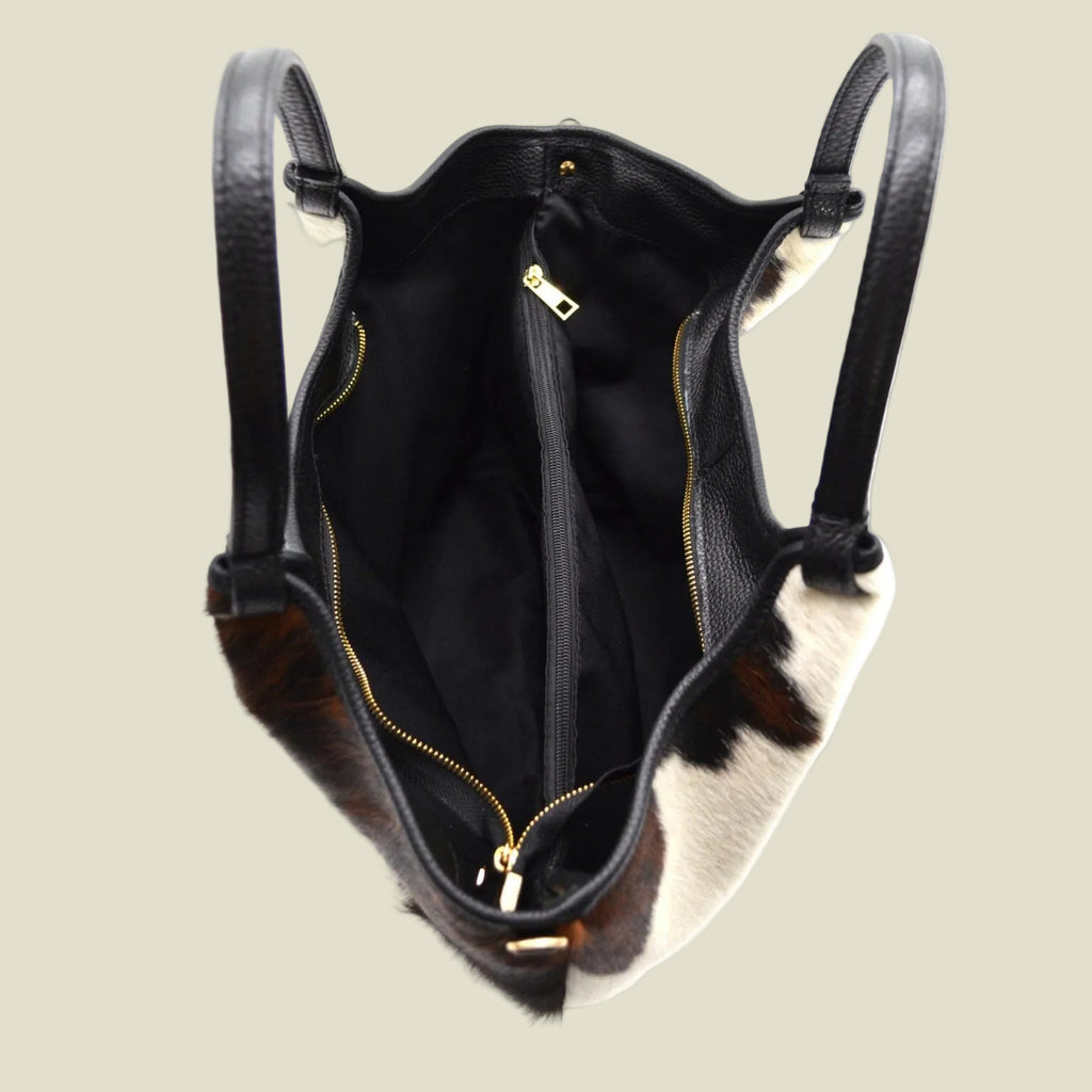 A cowhide shoulder bag shown from an overhead angle, featuring a black leather trim and handles, a black interior, and gold-tone zippers. The cowhide pattern displays a mix of dark brown, black, and cream-white patches.