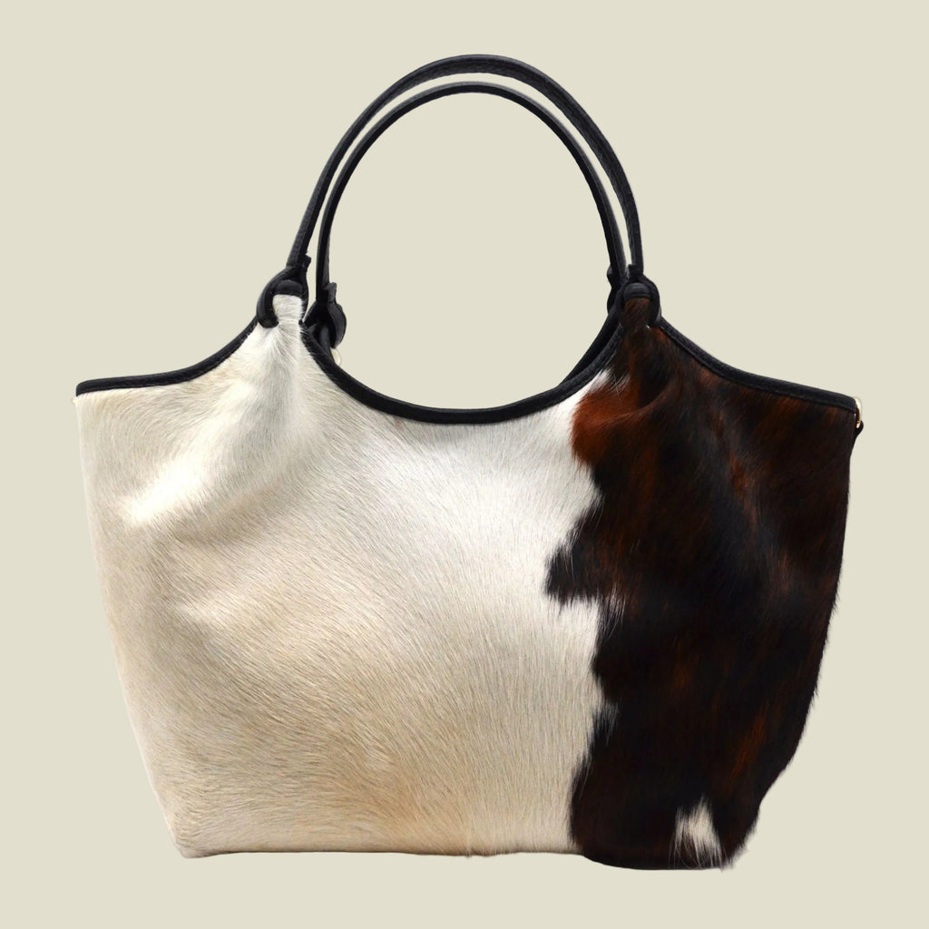 A cowhide shoulder bag with a split design featuring a white and brown hair-on-hide pattern on the front and a solid dark brown hide on the side. It has black leather handles and trim, photographed on a clean white background.